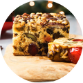 A thick slice of fruitcake on a light-colored wooden cutting board, topped with walnuts, pecans, candied red cherries, and golden raisins, with a blurred background of a red bowl and string lights.