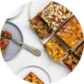 Overhead view of several slices of fruitcake with different toppings, including slivered almonds, candied red cherries, glazed orange peel, and other fruits and nuts, arranged on a white plate and surface with a silver fork resting on a gray plate in the upper left corner.