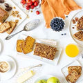 Overhead view of a breakfast table featuring sliced pumpkin bread, banana nut bread, a bowl of fresh blueberries, a bowl of cherries, two green apples, a glass of orange juice, and a small dish of coffee or dip, with an orange napkin, silver servingware, and additional bread slices in the background.