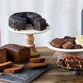 A dark chocolate bundt cake on a white and wood pedestal, a sliced chocolate loaf cake on a slate serving board, and a plate of assorted chocolates including squares and truffles, on a rustic wooden table with a partially visible bottle in the background.