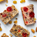 Three rectangular slices of traditional fruitcake with glossy red cherries, chopped walnuts, and golden raisins, arranged on a white surface with scattered nuts and dried fruit in the background.